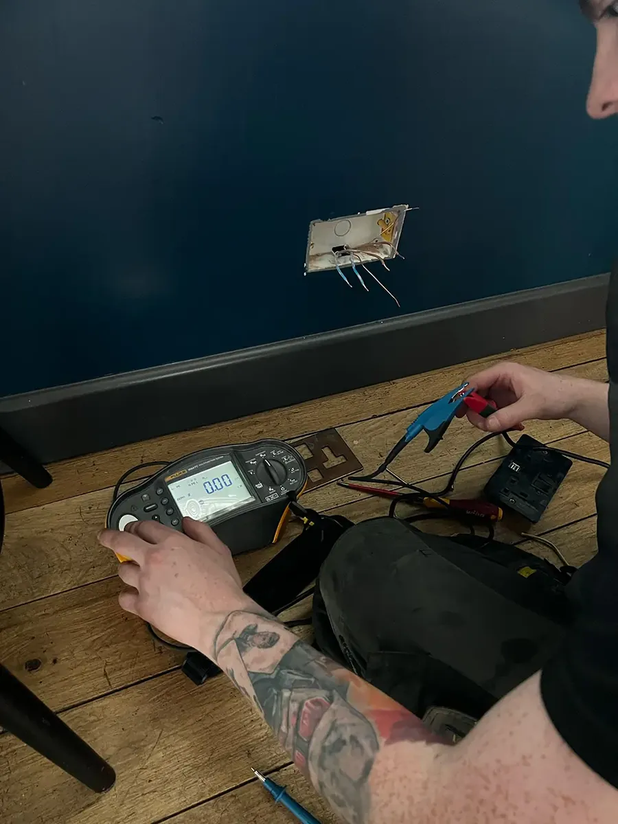 Jamie Grady, electrician, kneeling on a wooden floor testing a socket with a multifunction tester on a West Yorkshire Electrical job